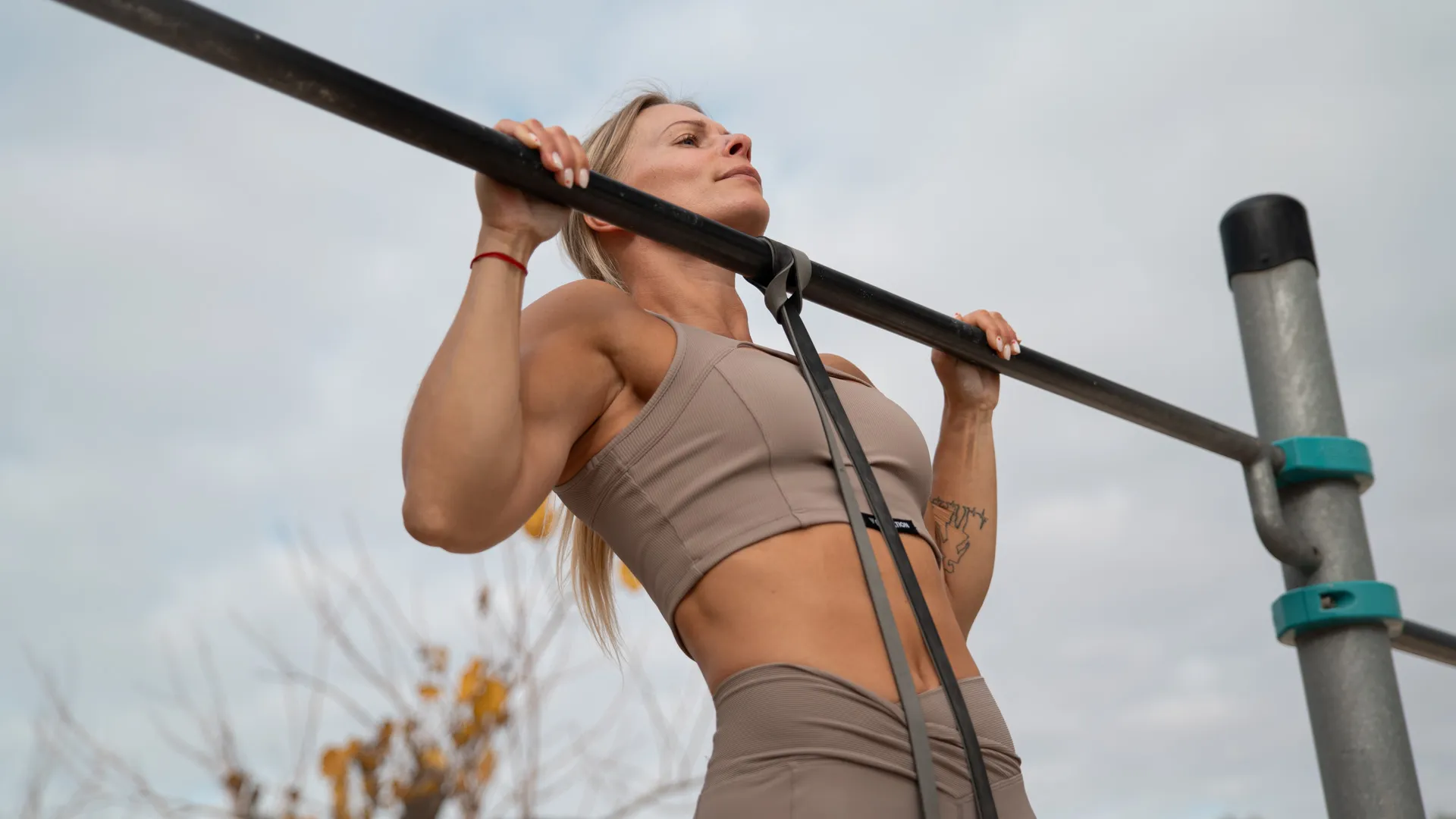 This image shows a person performing an assisted pull-up on an outdoor pull-up bar. A resistance band is attached to the bar and looped around the body to reduce the difficulty of the exercise by providing additional upward assistance. The individual is wearing a beige sports bra and leggings, emphasizing a strong and controlled posture. The focus is on the activation of the back, shoulders, and arm muscles during the upward pulling phase. This setup is ideal for building strength and progressing toward unassisted pull-ups.