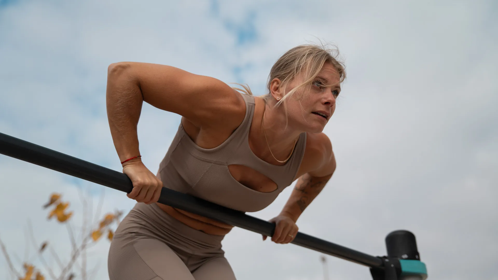 A woman in a beige workout outfit performing a dip on parallel bars outdoors, focusing intently and engaging her upper body muscles against a cloudy sky.