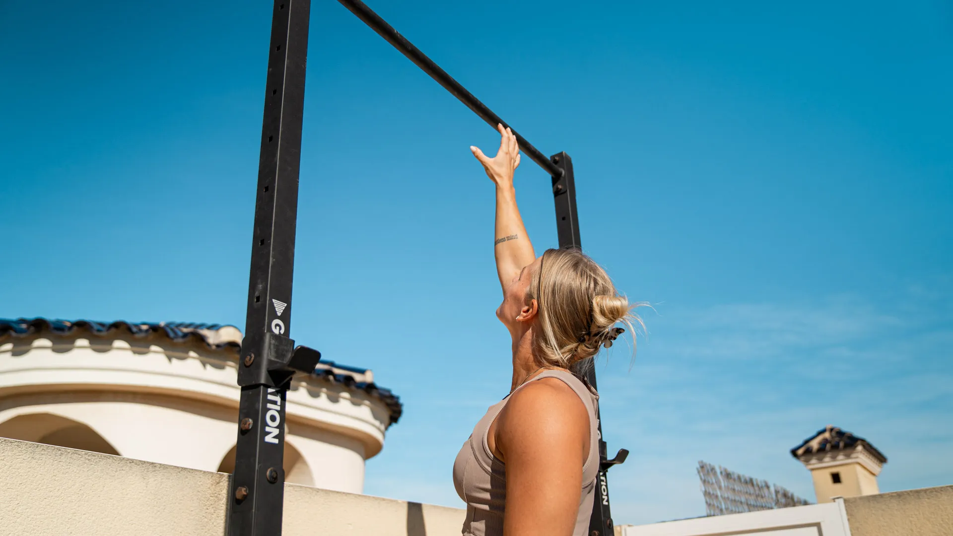 Woman reaching for the pull-up bar in an outdoor training environment, wearing a beige workout set, with clear blue skies and a rooftop in the background.